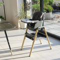 Lifestyle view of Nova Evolutionary Highchair in Grey/Oak placed beside a dining table, showcasing its stylish appearance and functional setup with tray and footrest in a home environment.