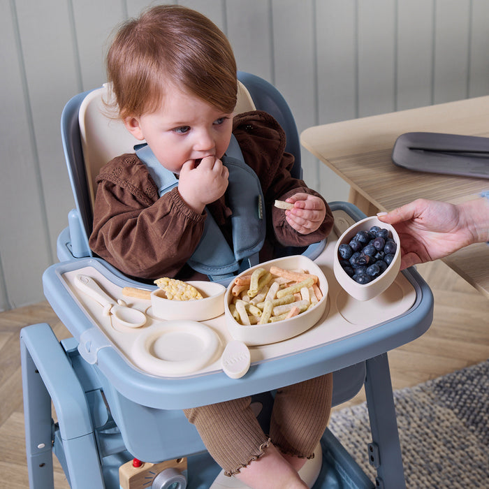 A child eating snacks while sitting in the Nova Maison highchair, with the accessory tableware set displayed. A bright lifestyle image showcasing independent mealtimes and the practical design of the Nova accessories.