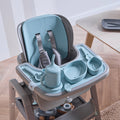 Nova Maison highchair with the accessory pack in blue, including a tray, sippy cup, three nesting bowls, fork and spoon, on a wooden floor.