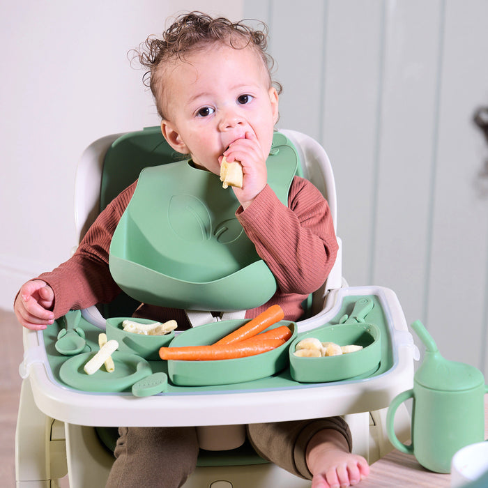 A baby sitting comfortably and eating in the Nova Maison highchair, with the green and white accessory pack showcased.