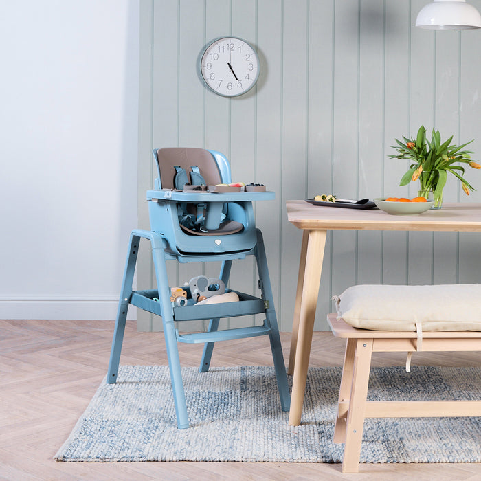 Nova Maison 7 in 1 highchair in blue with the accessory pack in a dinning setting with a table and chair and a clock on the wall.