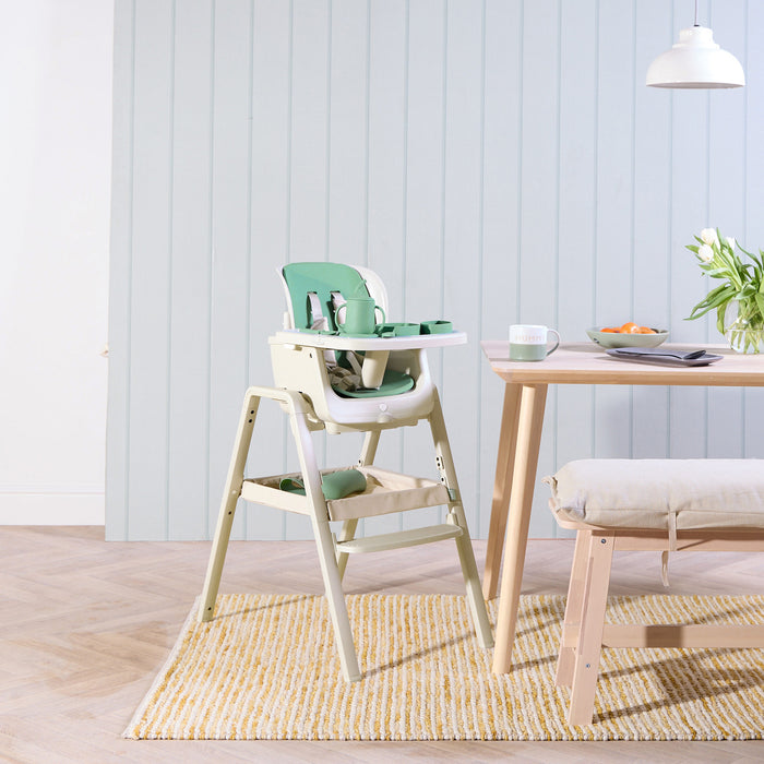 Nova Maison highchair in beige with a green accessory pack next to a wooden dining table with a light blue wall in the background