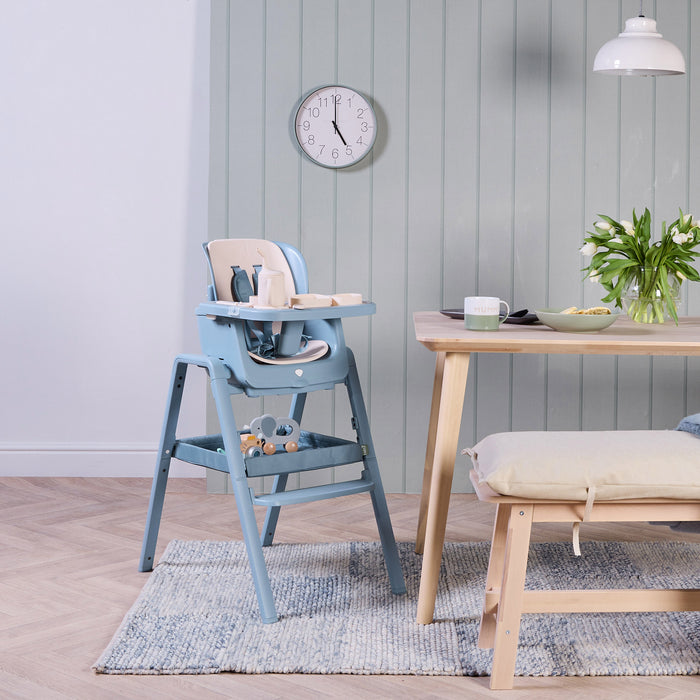 Nova Maison highchair in blue with a beige accessory pack next to a wooden dining table with a light blue wall in the background