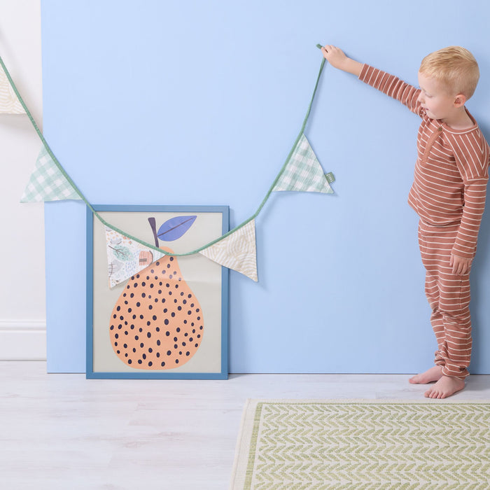 Toddler holding good life nursery bunting against blue wall with framed pear artwork and green patterned rug.