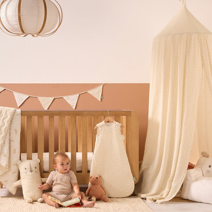 Baby sitting in front of a cot bed with bedding, toys, and canopy from the Love Cats collection in a nursery setting.