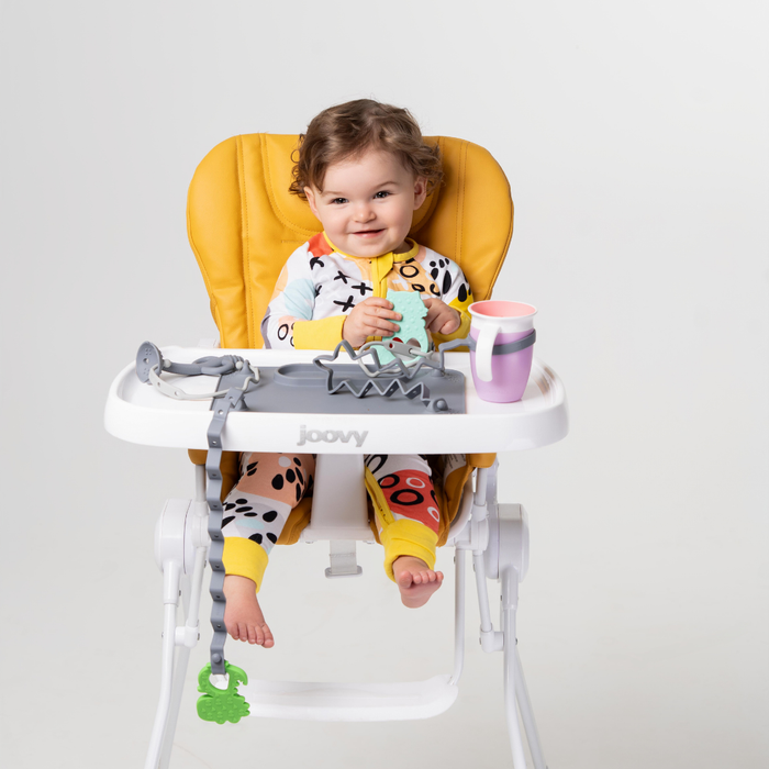 baby sitting in high chair with busy baby suction placemat and toy bungee holding dino teether