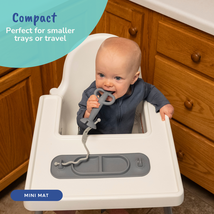 baby sitting in high chair chewing on 2-in-1 teether and training spoon attached with toy bungee to the pewter busy baby suction mini placemat