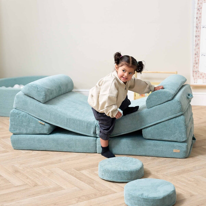Smiling toddler climbing on the Playrama Montessori Kids Play Sofa in ocean stone, set up as a slope for active play and movement