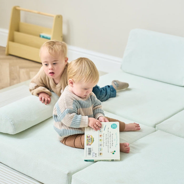 Two toddlers sit on the Playrama Montessori kids play sofa in sea mist, one reading a book while the other leans on the cushion