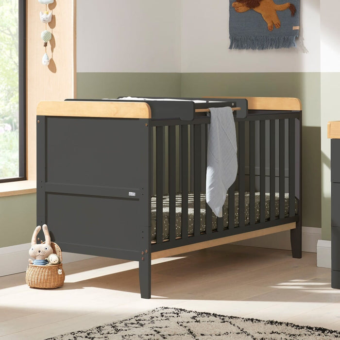 Rio Cot Bed and top changer in slate grey and honey oak styled in nursery setting, showcasing versatile function and modern tones.