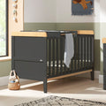Rio Cot Bed and top changer in slate grey and honey oak styled in nursery setting, showcasing versatile function and modern tones.