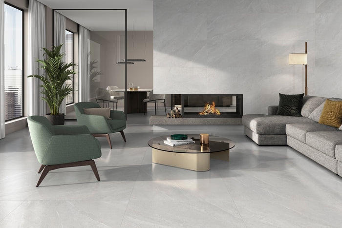 A modern living room with light gray stone-look porcelain floor tiles, featuring a gray sectional sofa, two green accent chairs, a round coffee table, and a built-in fireplace against a textured gray wall.