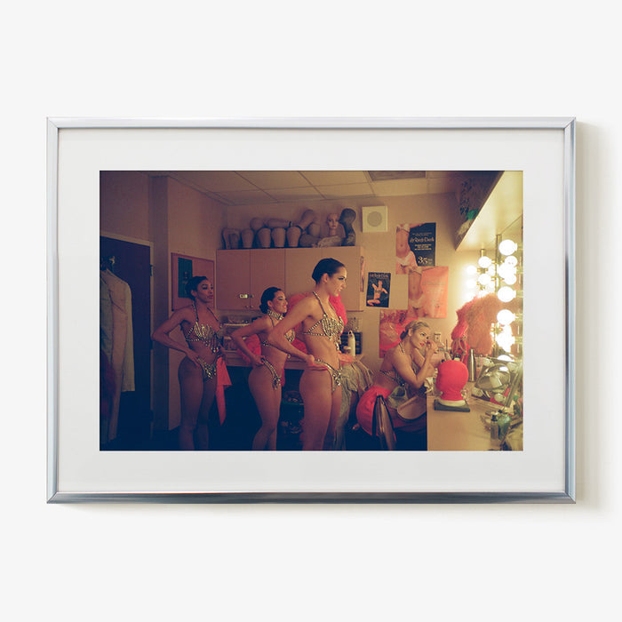 three performers in shimmering costumes and one applying makeup in a backstage dressing room in a silver frame