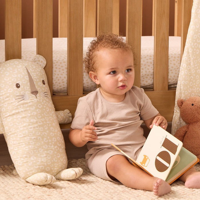 Toddler sitting beside the Soft Leopard Cuddle Cushion, reading a book on a textured rug in a calm neutral nursery setting