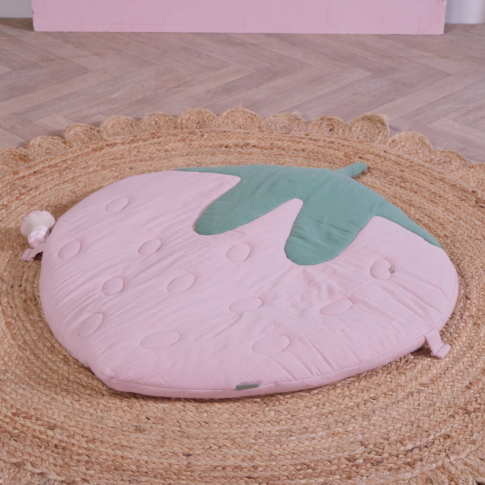 Strawberry-shaped padded baby playmat in soft pink muslin with green leaf detail, styled on a woven jute rug.