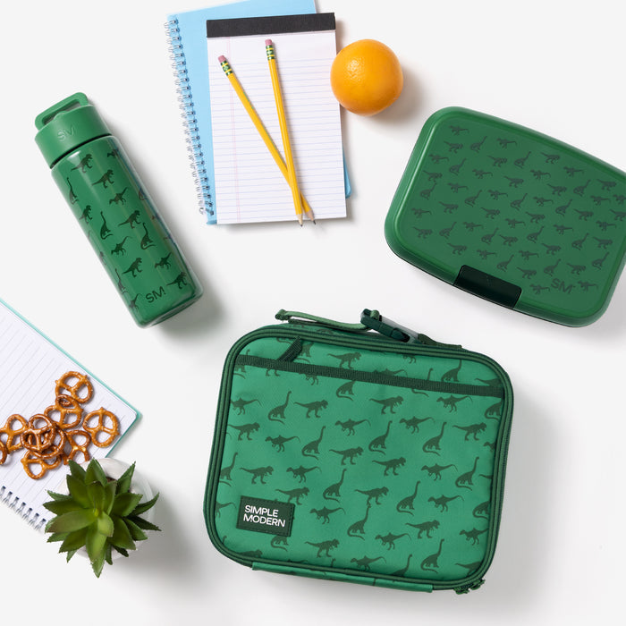 Simple Modern kids' collection featuring kelly green insulated water bottle, lunch container, and carrying case with dinosaur print, arranged with school supplies including yellow pencils, blue notebook, orange, and pretzel