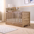 Lifestyle view of the Modena cot bed in oak with changing top attached and spacious underbed storage drawer for practical nursery use