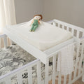 A white Universal Cot Top Changer placed on top of a cot with white bed rails and a patterned bedsheet. A stuffed toy is seated on the mattress.