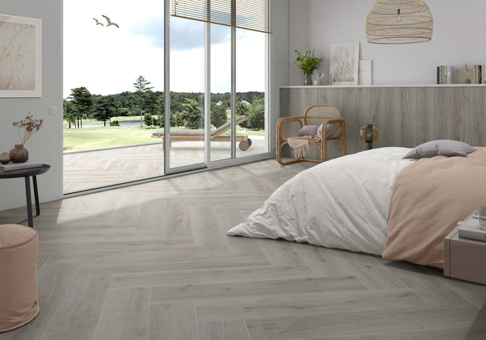 Light gray herringbone wood-look tile flooring shown in a modern, sunlit bedroom with a bed, large windows, and an outdoor view.