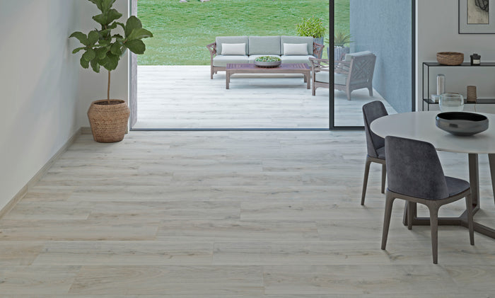 Light-colored wood-look tile flooring installed in a modern interior room, with a dining table and chairs on the right and an open doorway leading to a patio and green lawn.