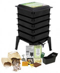 The Natures Footprint Worm Factory 365 Vermicomposting System, in black with multiple tiers and legs, comes with a starter kit including paper scraps, a handbook, a small bin, and composting tools displayed in front.