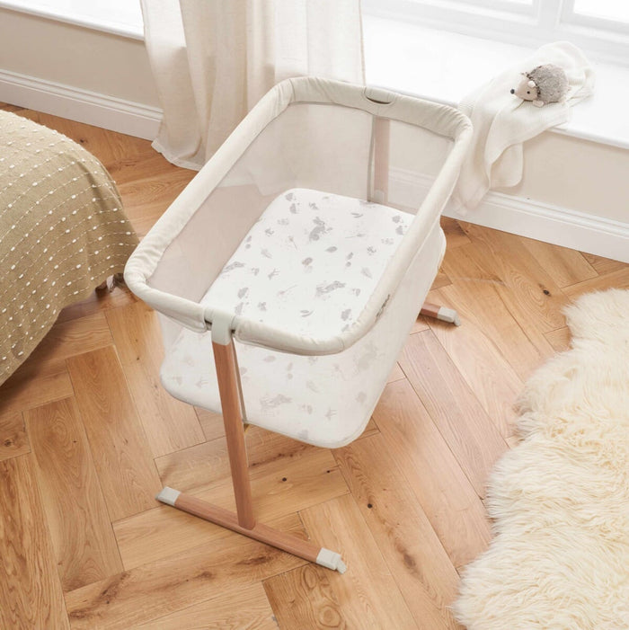 Top view of ZiZee Essential Crib in Oak and Ecru colours, featuring a mesh surrounding, a white mattress, and wooden legs, placed next to a fluffy white rug.