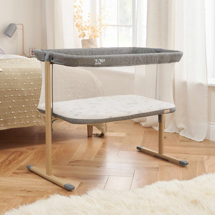 ZiZee Essential Crib in Oak and Charcoal colours, featuring a mesh surrounding, a white mattress, and wooden legs, placed next to a fluffy white rug.